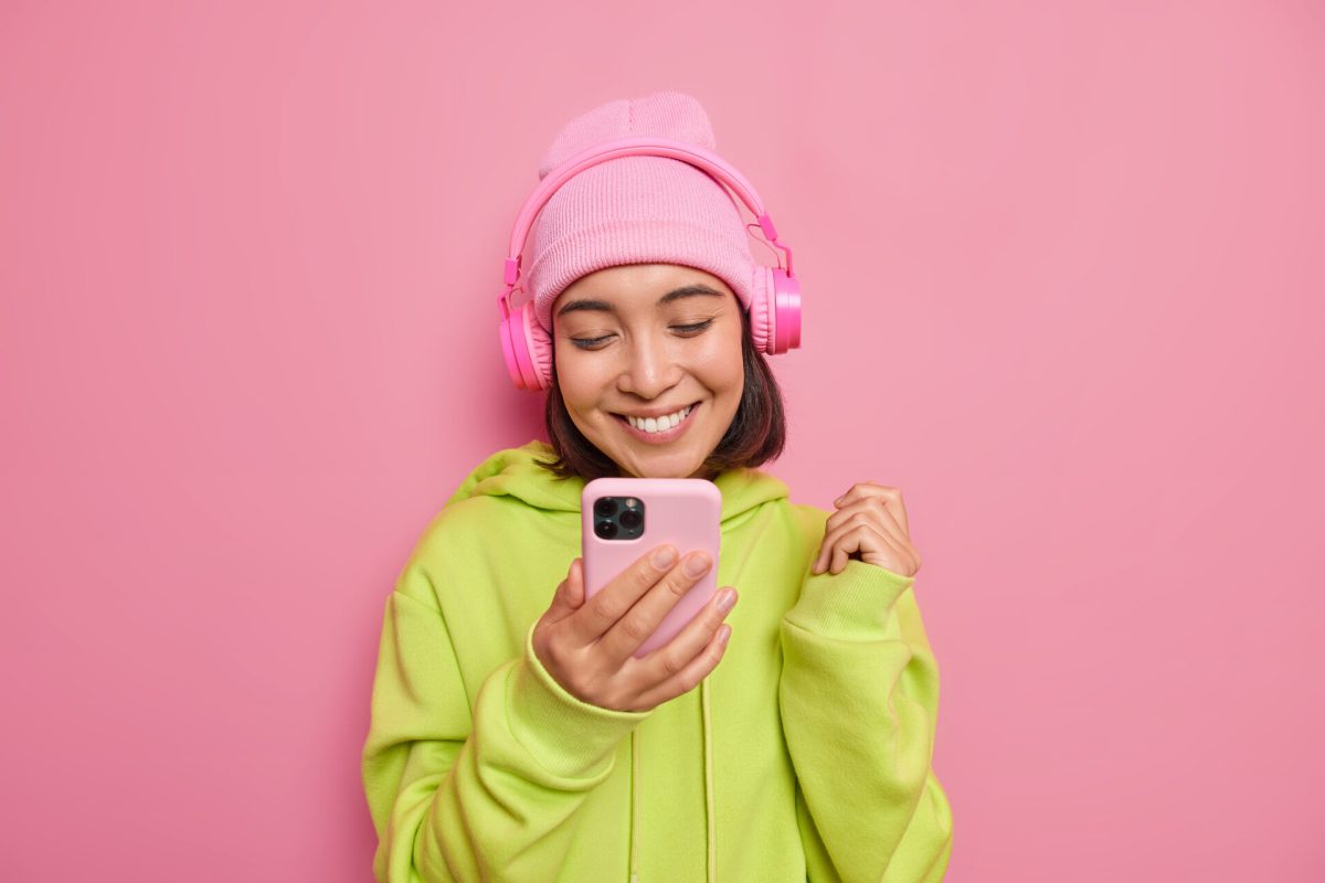 Beautiful glad Asian female teenager looks happily at smartphone listens music via headphones enjoys favorite playlist wears hat and green sweatshirt isolated over pink background. Technology