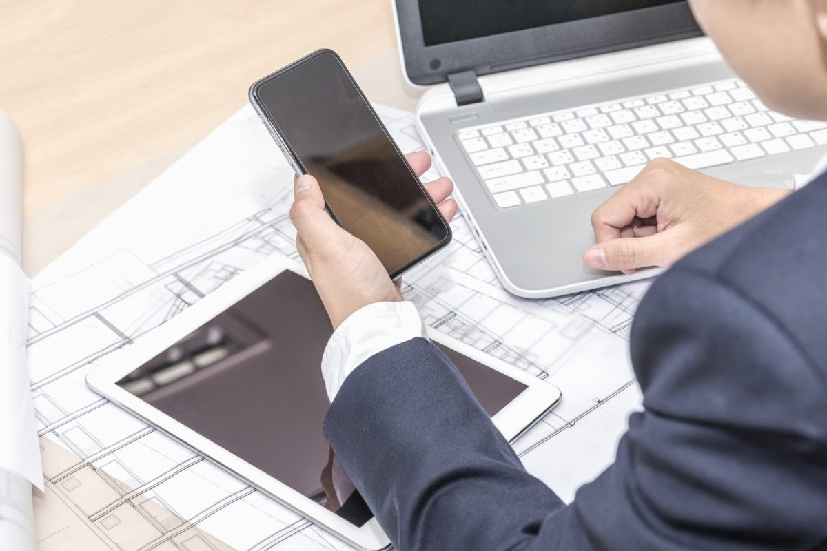 close up of businessman holding smartphone with black screen over tablet pc computer notebook blueprints paper and blueprint rolls on the table in the office, architecture contractor developer and renovation concept