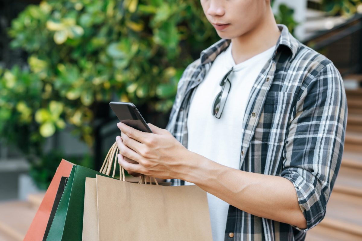 Close up, Young handsome man holding paper bag and use smartphone on stair of shopping mall, Lifestyle and new normal concept, copy space