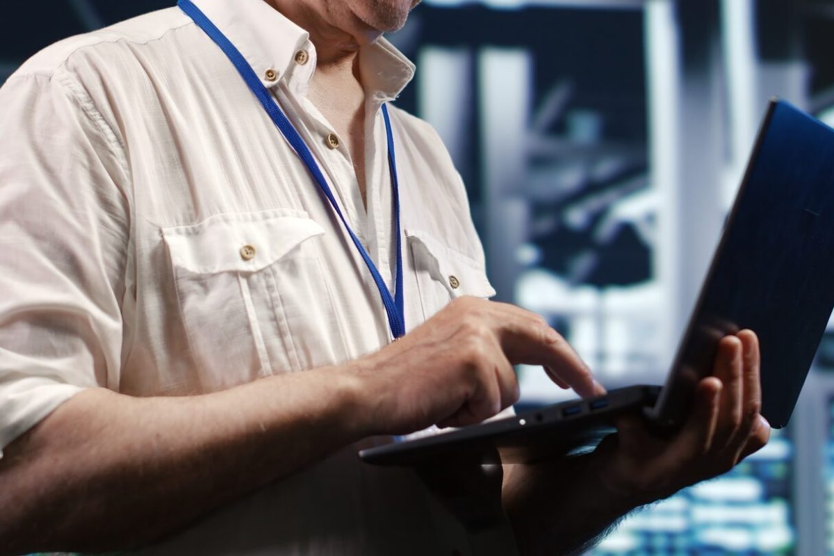 Experienced IT professional navigating network of mainframes in industrial server hub. Employee with laptop ensuring smooth operations, doing optimizations and maintenance in data center, close up