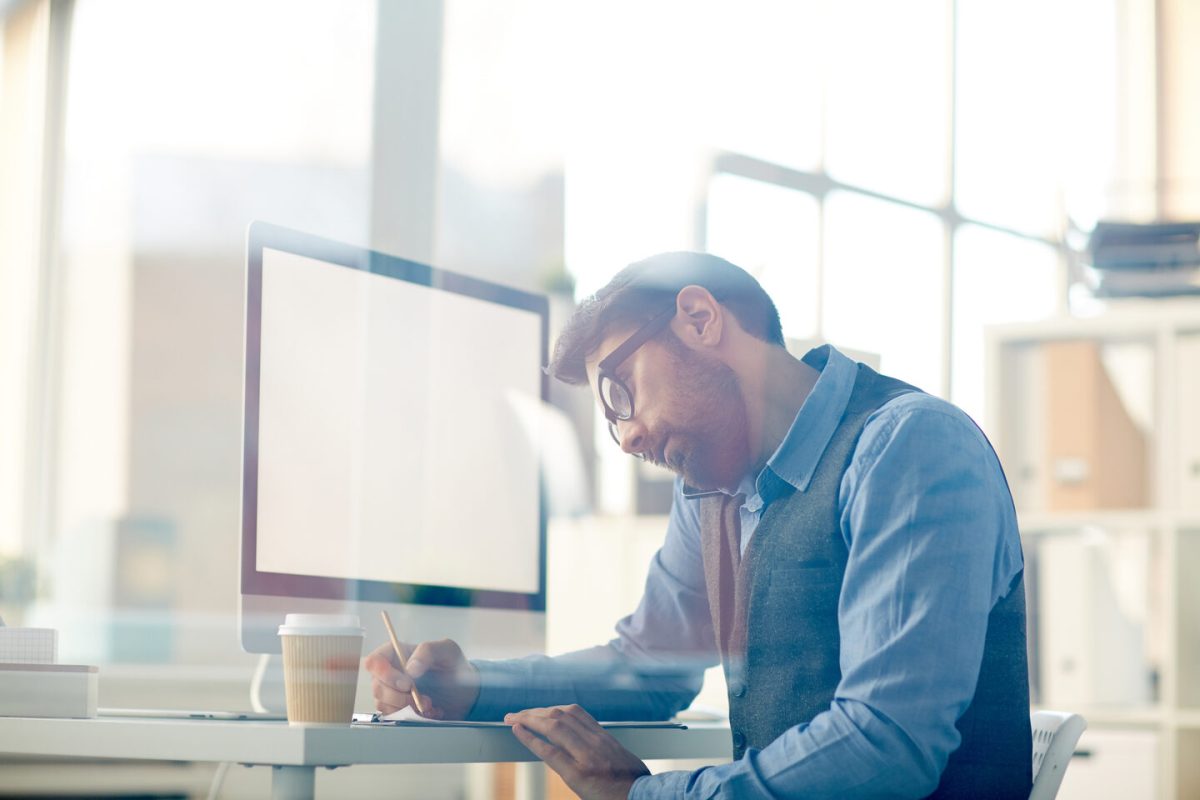 Young man talking on smartphone and making notes on paper while sitting by his workplace in office