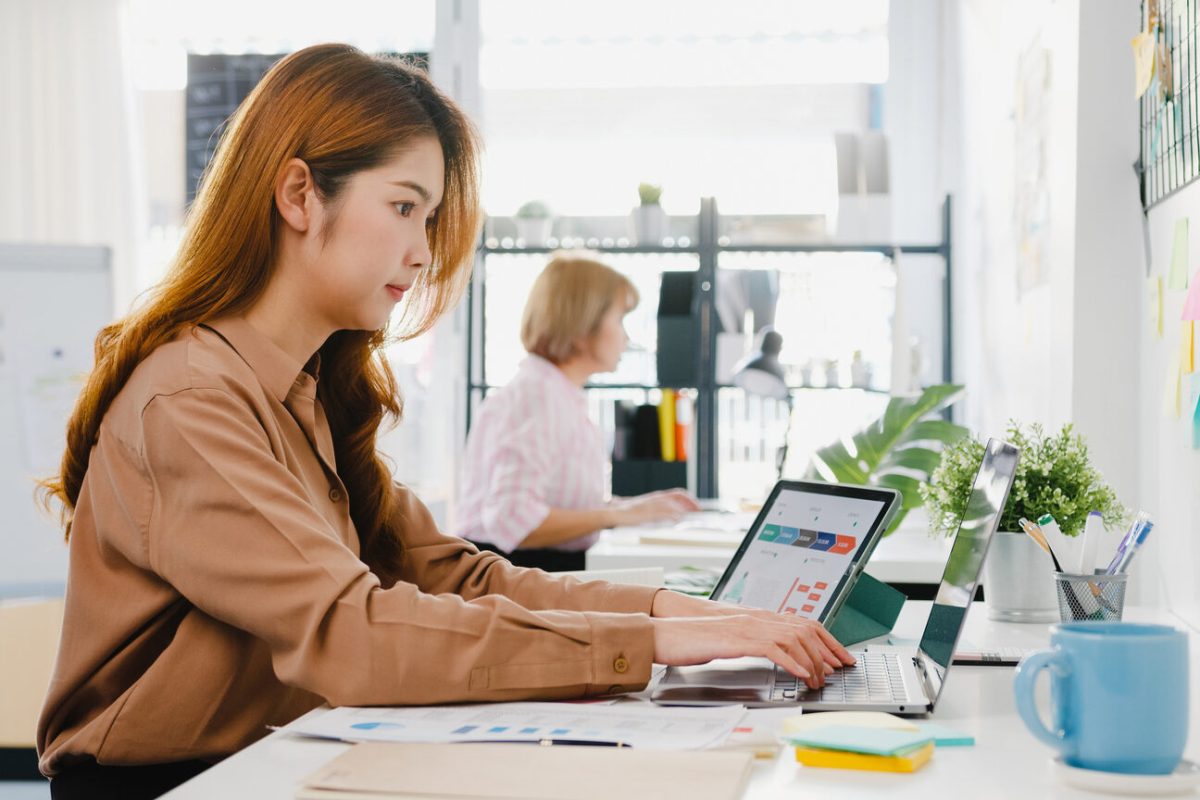 Young Asia businesswoman entrepreneur social distancing in new normal situation for virus prevention while using laptop computer and tablet back at work in office. Lifestyle after coronavirus.